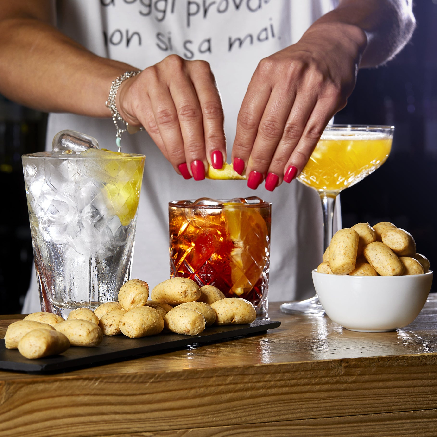 Hands squeezing lemon over a cocktail with I Macoritti mini breadsticks served on a board and in a bowl alongside mixed drinks